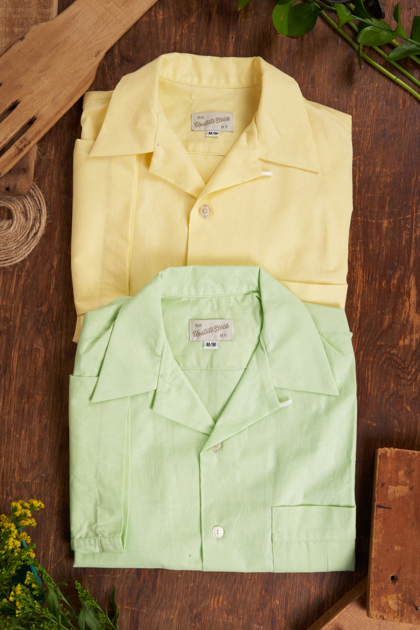 Two folded shirts, one yellow and one green, on a wooden surface with plants around.