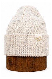 An off-white knit beanie hat with subtle multicolor flecks, displayed on a rustic brown wooden stand against a clean white background. A small tan leather patch on the side cuff features the brand name 'The Unstitched Stitch'.