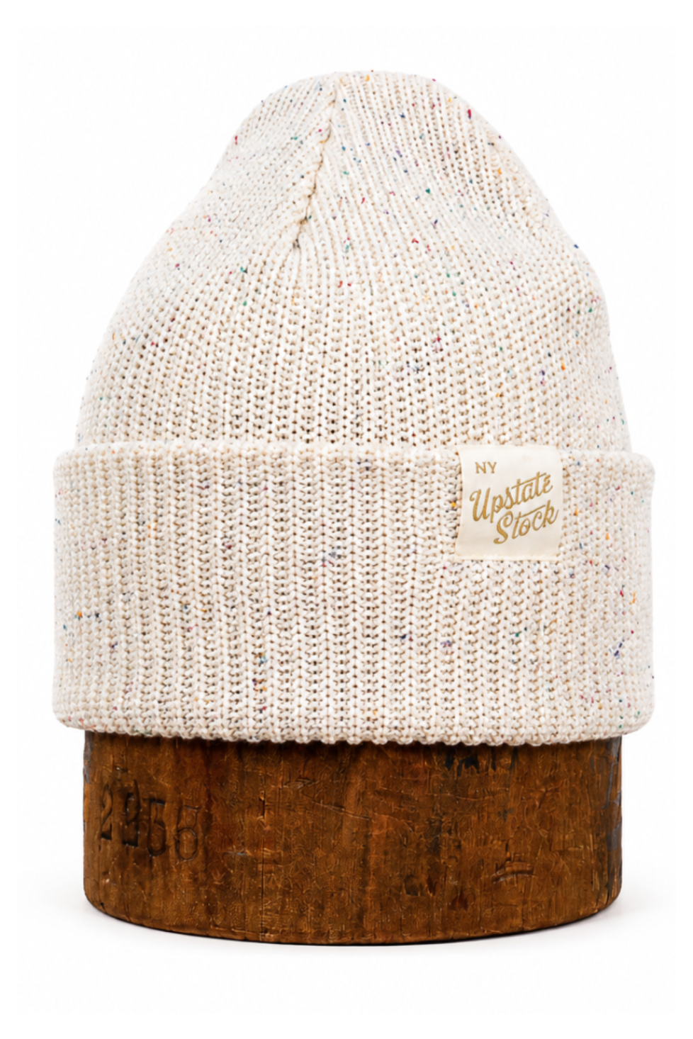 An off-white knit beanie hat with subtle multicolor flecks, displayed on a rustic brown wooden stand against a clean white background. A small tan leather patch on the side cuff features the brand name 'The Unstitched Stitch'.