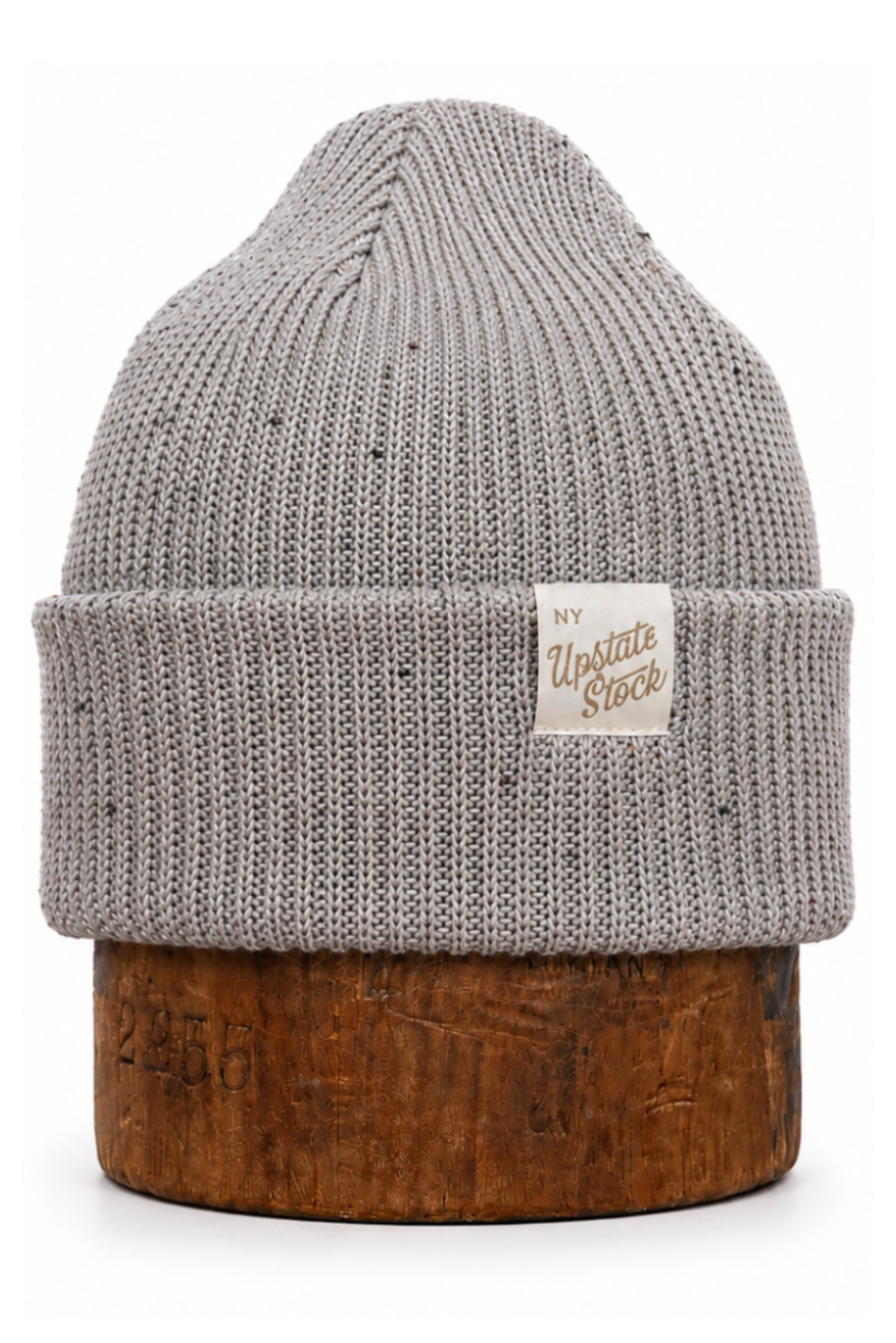 A light gray speckled knit beanie hat with a folded cuff and a small white brand patch reading 'Upstate Stock', displayed on a brown wooden stand against a white background.