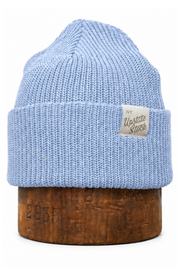 A light blue ribbed knit beanie hat displayed on a round wooden stand against a plain white background. A small beige fabric label is stitched onto the folded cuff, and the wooden base has faint embossed numbers.