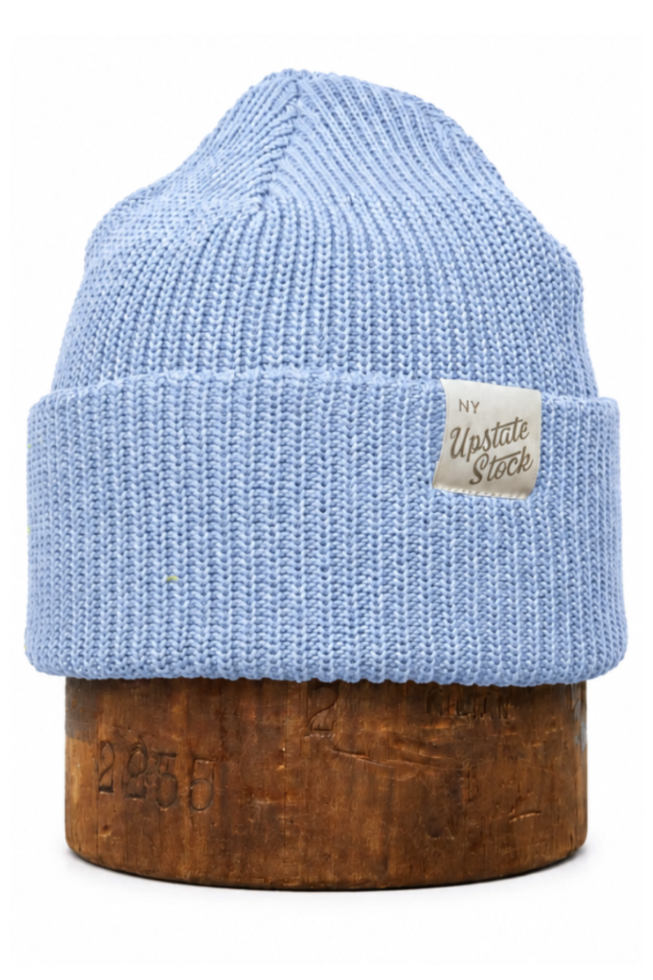 A light blue ribbed knit beanie hat displayed on a round wooden stand against a plain white background. A small beige fabric label is stitched onto the folded cuff, and the wooden base has faint embossed numbers.