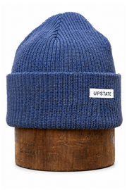 A blue ribbed knit beanie hat with a folded cuff and a small white rectangular label reading 'UPSTATE' on the side, displayed on a round wooden stand against a plain white background.