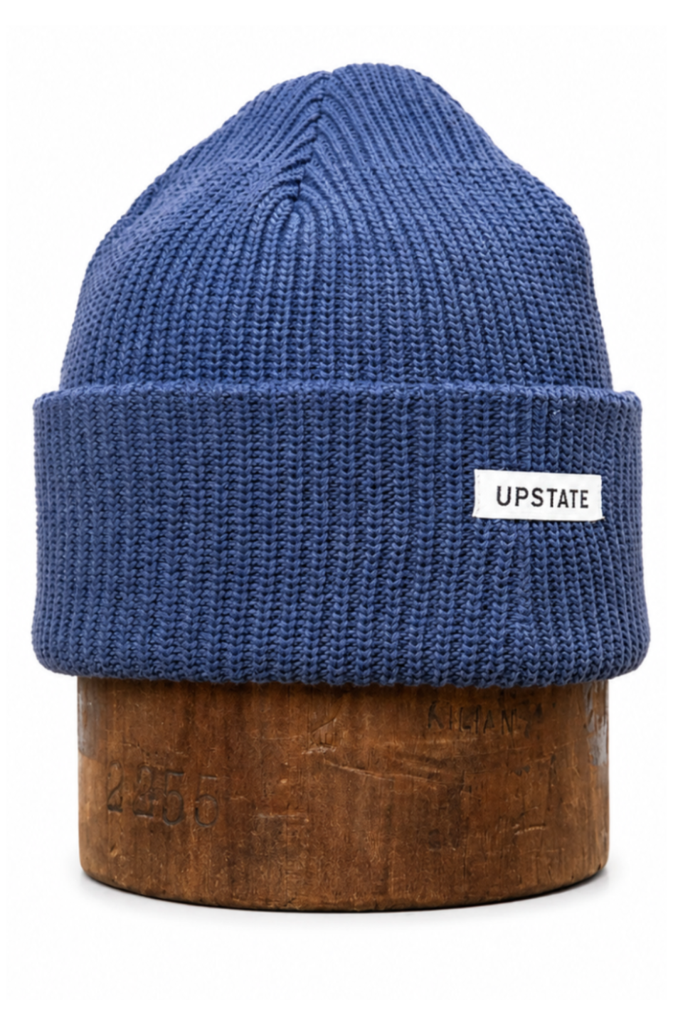 A blue ribbed knit beanie hat with a folded cuff and a small white rectangular label reading 'UPSTATE' on the side, displayed on a round wooden stand against a plain white background.