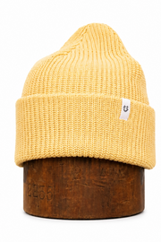 A mustard yellow ribbed knit beanie hat displayed on a dark brown wooden stand against a plain white background.