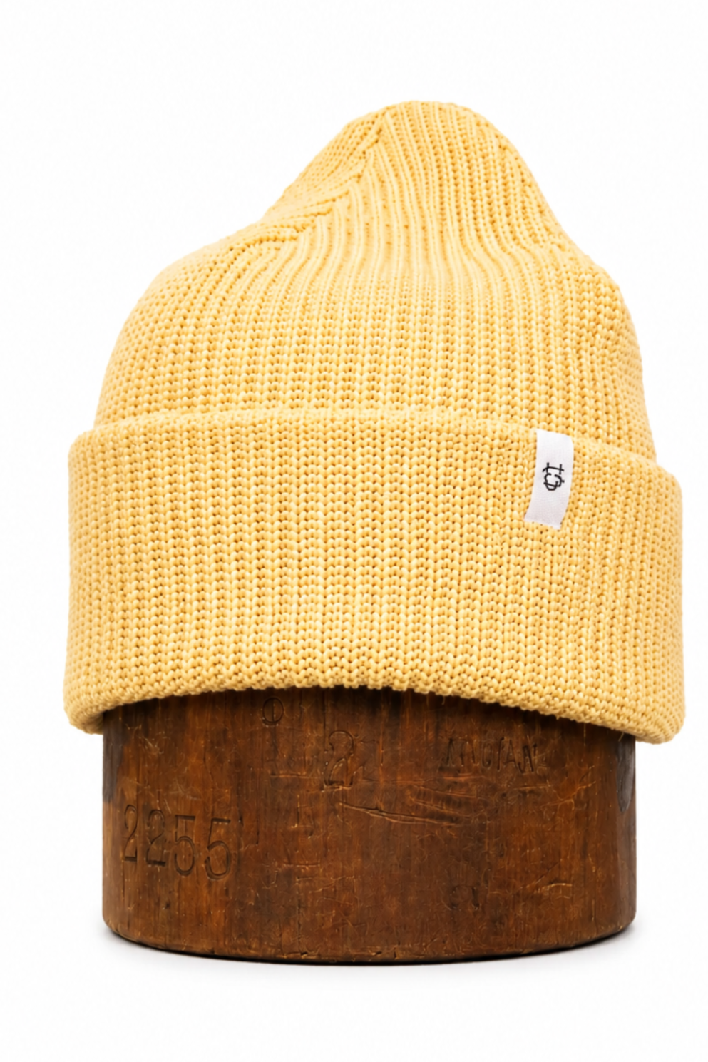 A mustard yellow ribbed knit beanie hat displayed on a dark brown wooden stand against a plain white background.