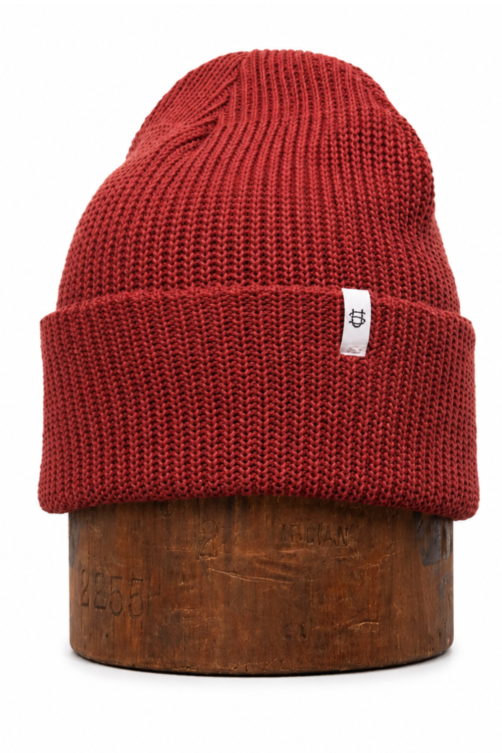 A ribbed knit beanie hat in a deep red or burgundy color, folded over at the brim with a small white brand tag on the side, displayed on a wooden stand against a plain white background.