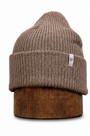 A ribbed knit beanie hat in taupe brown, displayed on a wooden stand against a plain white background. A small white square tag with a black logo is visible on the side of the cuff.