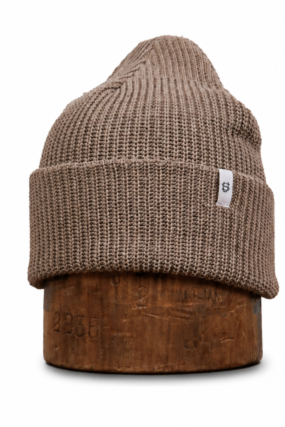 A ribbed knit beanie hat in taupe brown, displayed on a wooden stand against a plain white background. A small white square tag with a black logo is visible on the side of the cuff.