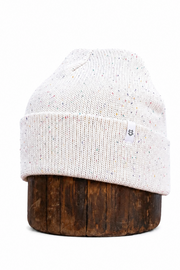 A cream-colored knit beanie hat with subtle multicolor flecks, displayed on a dark brown wooden stand against a solid white background.