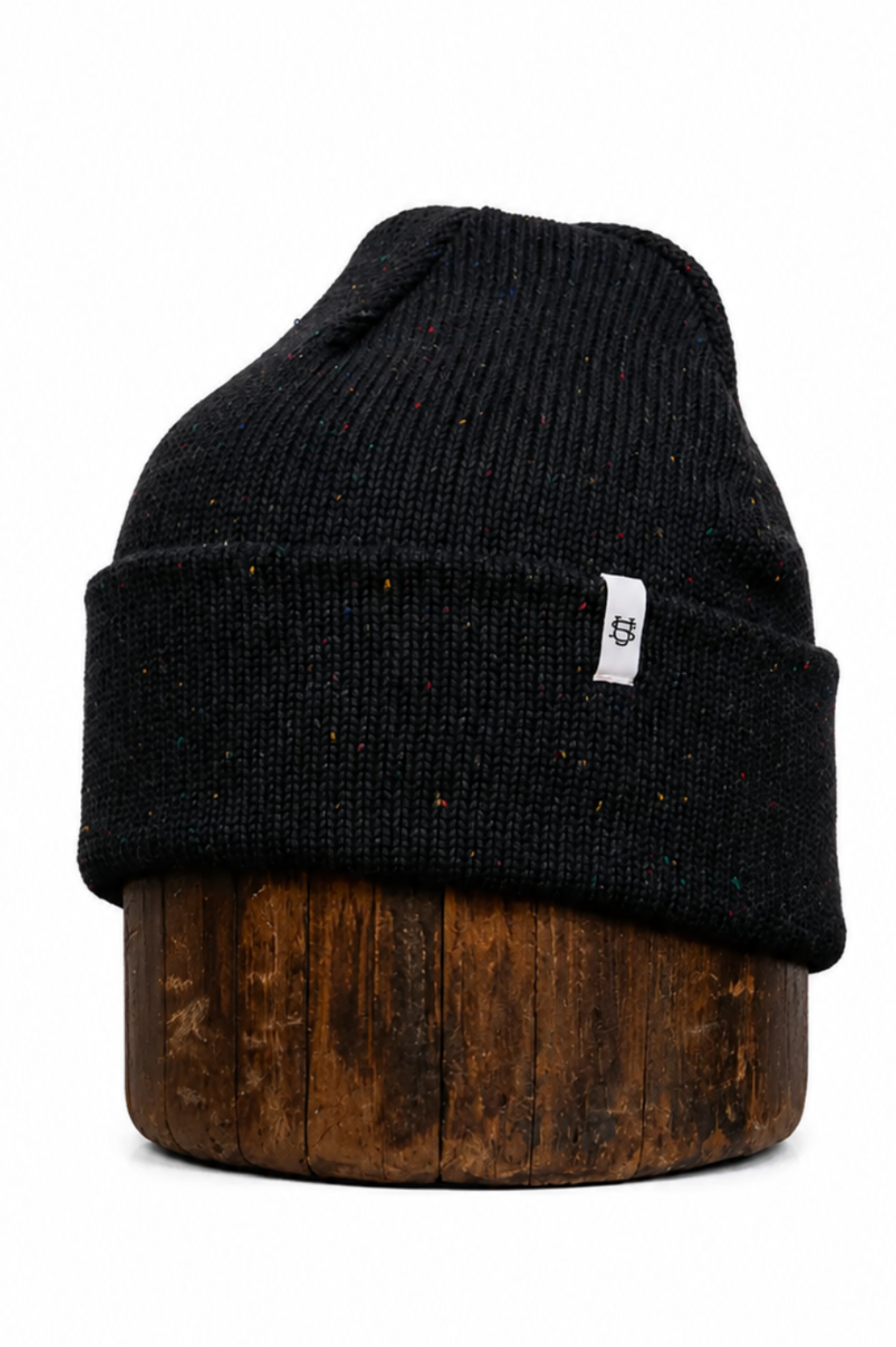 A black knit beanie hat with subtle speckles, displayed on a dark wood block against a solid white background. A small white square tag is visible on the side cuff.