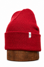 A bright red ribbed knit beanie hat displayed on a round wooden stand against a plain white background.