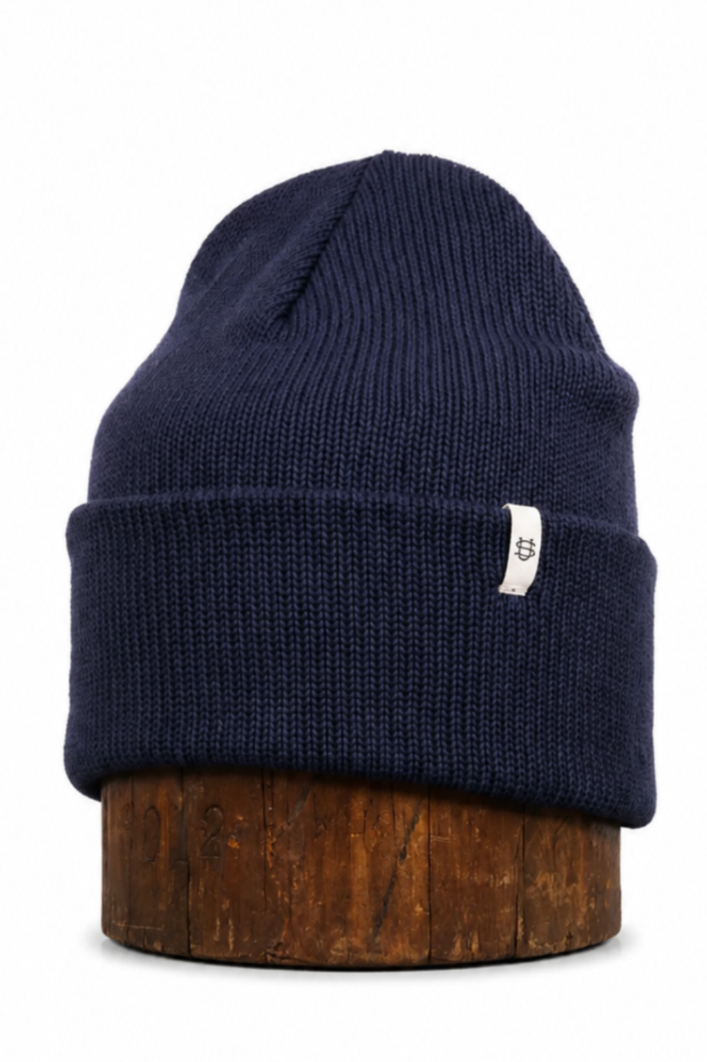 A navy blue ribbed knit beanie hat with a small white tag on the side, displayed on a brown wooden stand against a plain white background.