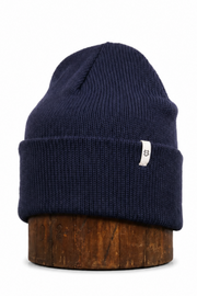 A navy blue ribbed knit beanie hat with a small white tag on the side, displayed on a brown wooden stand against a plain white background.