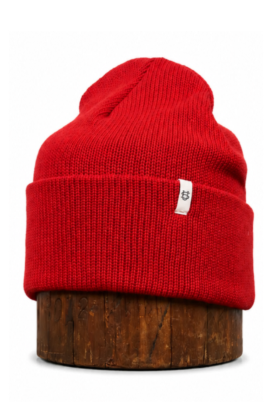 A bright red ribbed knit beanie hat displayed on a round wooden stand against a plain white background.