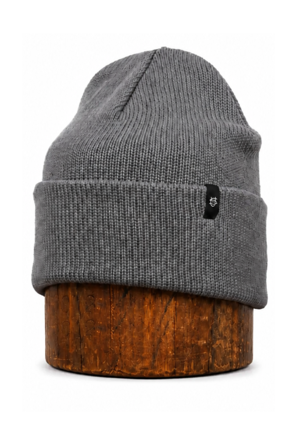 A gray ribbed knit beanie hat with a small black logo patch on the front cuff, displayed on a round wooden stand against a plain white background.