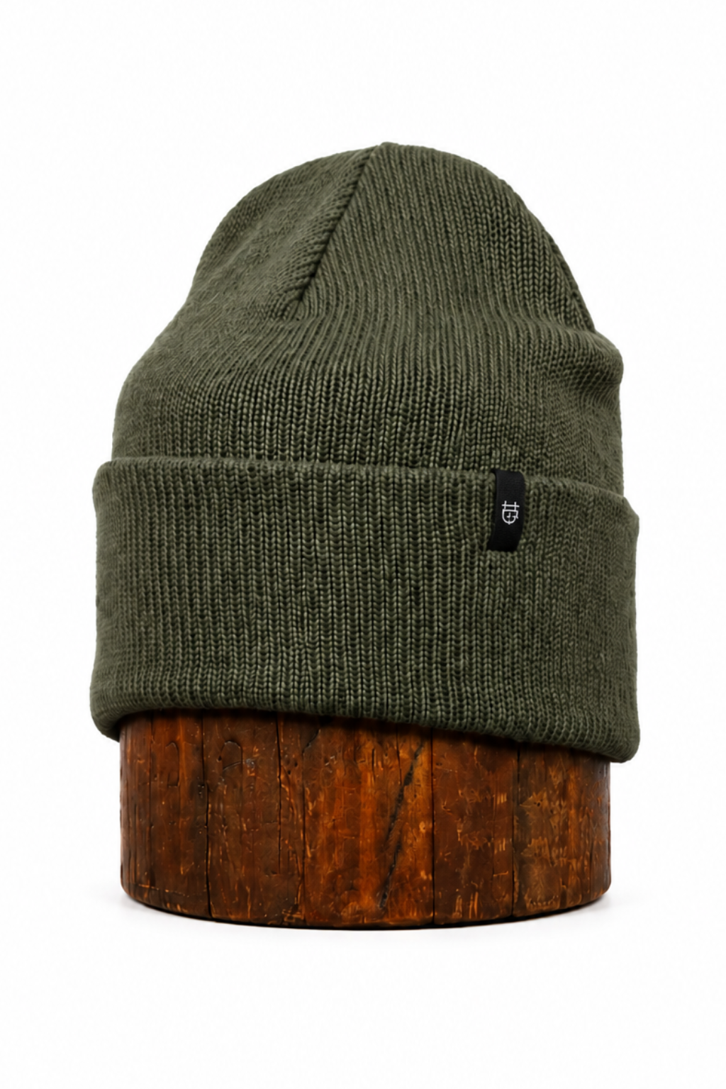 An olive green ribbed knit beanie hat with a small black logo patch on the front cuff, displayed on a round wooden stand against a plain white background.