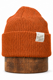An orange knit beanie hat with a folded cuff and a small white brand label, displayed on a wooden stand against a white background.