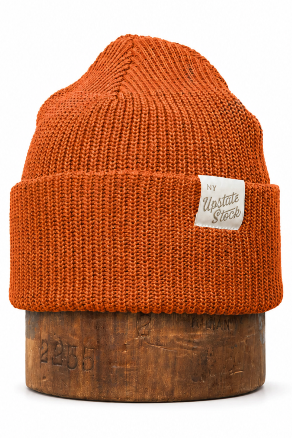 An orange knit beanie hat with a folded cuff and a small white brand label, displayed on a wooden stand against a white background.