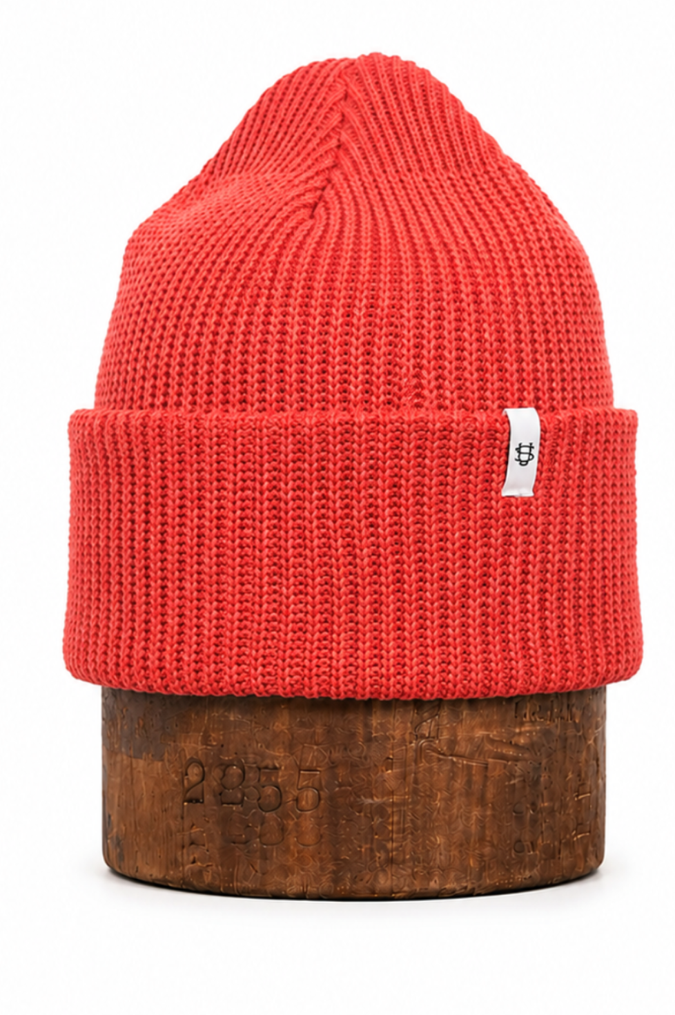 A bright red ribbed knit beanie hat displayed on a round wooden block against a plain white background.