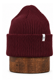 A ribbed knit beanie hat in deep burgundy, displayed on a brown wooden stand against a plain white background. A small white square tag is attached to the folded cuff.