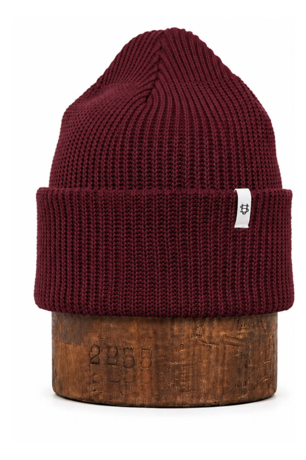 A ribbed knit beanie hat in deep burgundy, displayed on a brown wooden stand against a plain white background. A small white square tag is attached to the folded cuff.