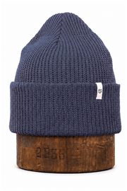 A navy blue ribbed knit beanie hat with a small white tag, displayed on a wooden block against a white background.