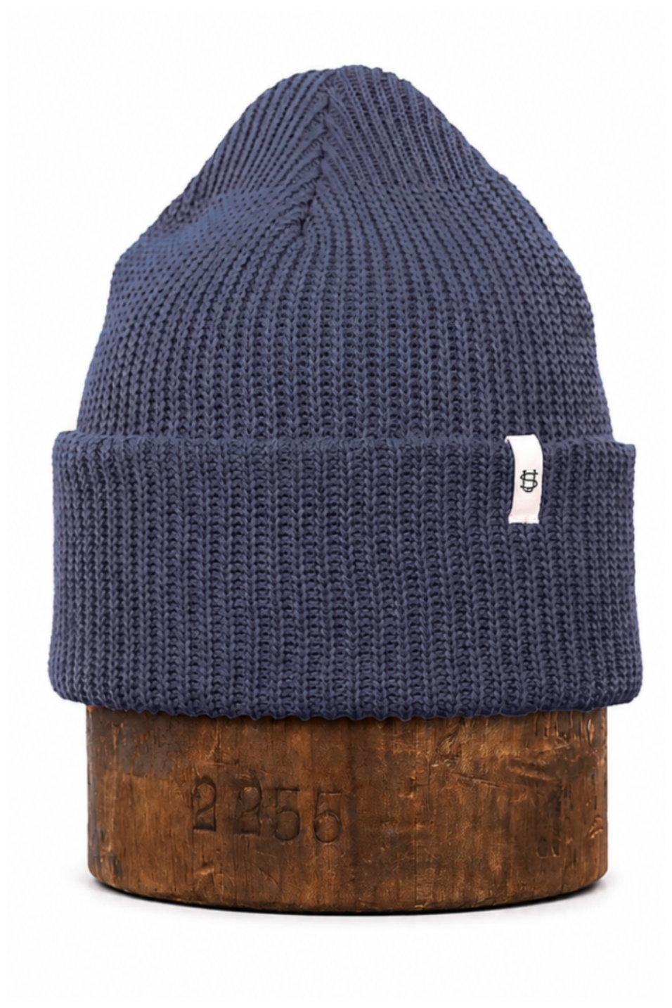 A navy blue ribbed knit beanie hat with a small white tag, displayed on a wooden block against a white background.