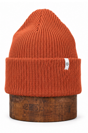 An orange ribbed knit beanie hat displayed on a wooden stand against a plain white background.