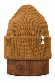 A ribbed knit beanie hat in a warm camel brown color, displayed on a rustic wooden block against a clean white background.