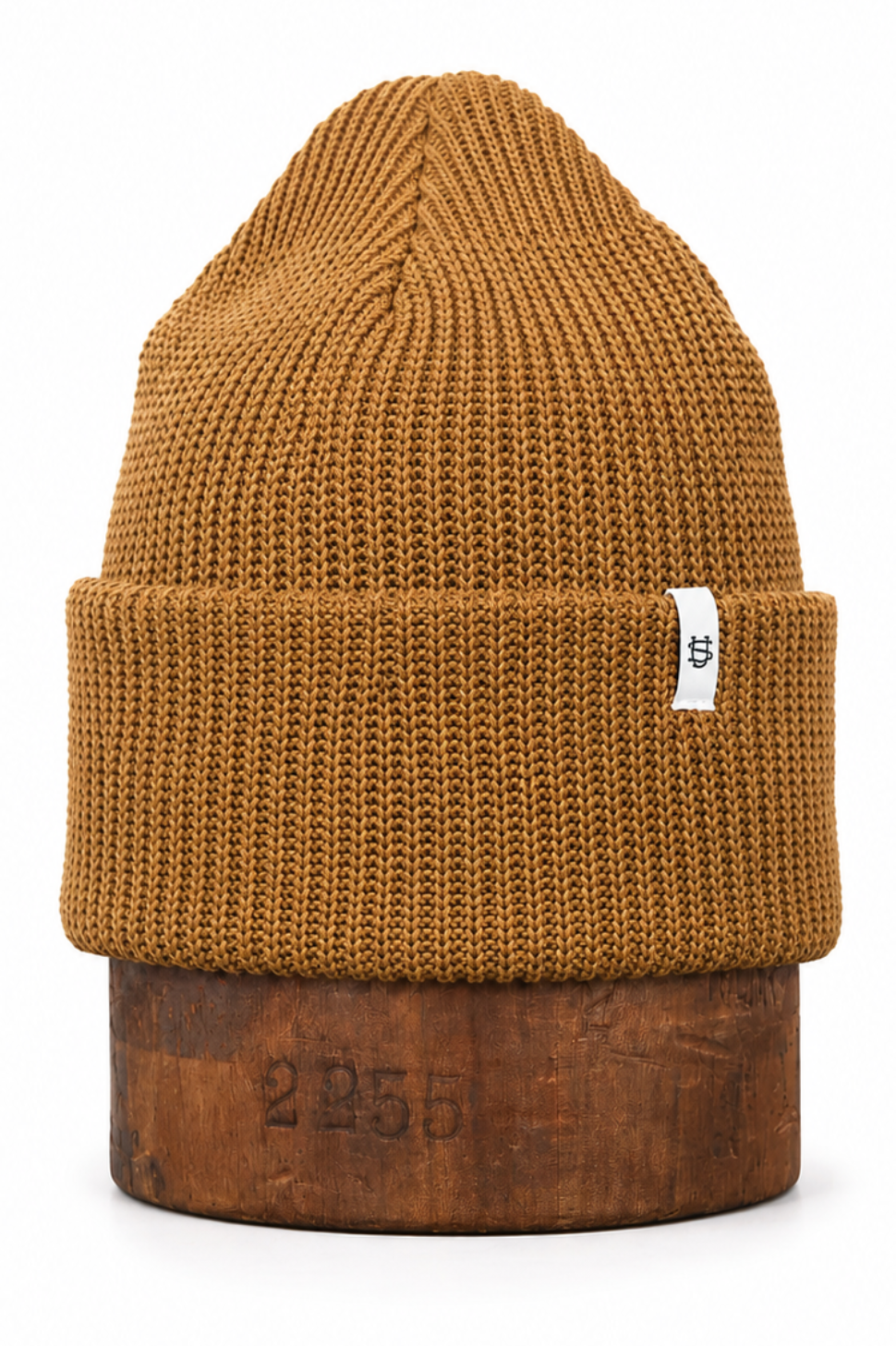 A ribbed knit beanie hat in a warm camel brown color, displayed on a rustic wooden block against a clean white background.