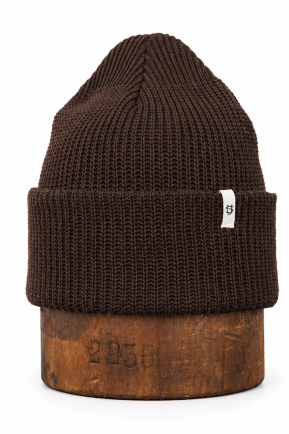 A dark brown ribbed knit beanie hat with a small white tag on the side, displayed on a rustic wooden block against a solid white background.