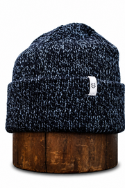 A navy blue and black marled knit beanie hat with a white brand tag, displayed on a dark wood block against a white background.