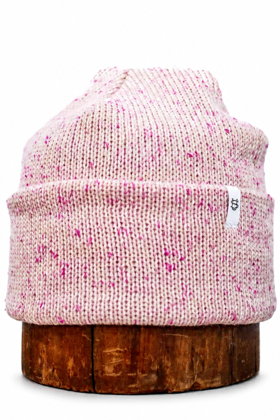 A light pink and white speckled knit beanie hat displayed on a rustic wooden stand against a plain white background.