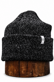 A dark gray and black heathered knit beanie hat with a small white logo patch on the side, displayed on a wooden stand against a white background.