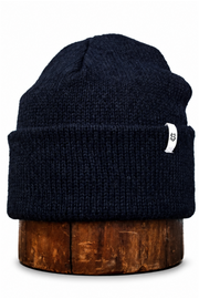 A dark navy blue ribbed knit beanie hat with a small white side tag, displayed on a round wooden stand against a plain white background.
