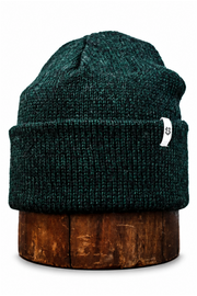 A dark green heathered knit beanie hat with a small white tag, displayed on a round wooden stand against a plain white background.