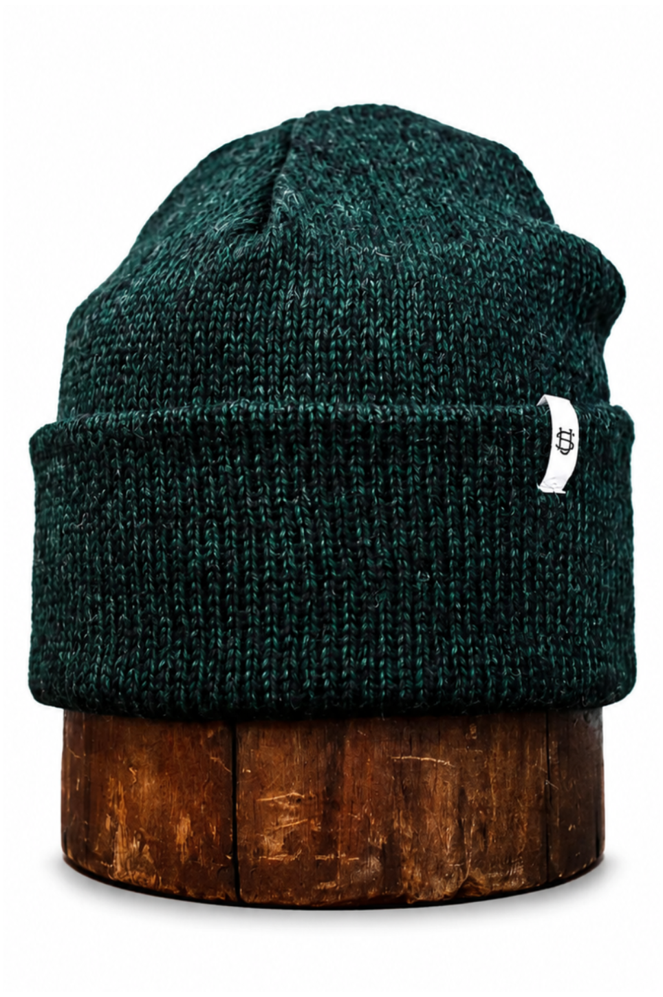A dark green heathered knit beanie hat with a small white tag, displayed on a round wooden stand against a plain white background.