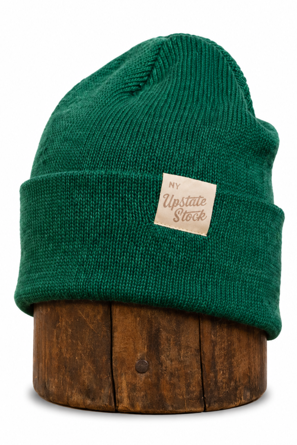 A dark green ribbed knit beanie hat with a folded cuff, displayed on a round wooden stand against a plain white background. A small beige fabric label on the side reads 'NY Upstate Stock'.