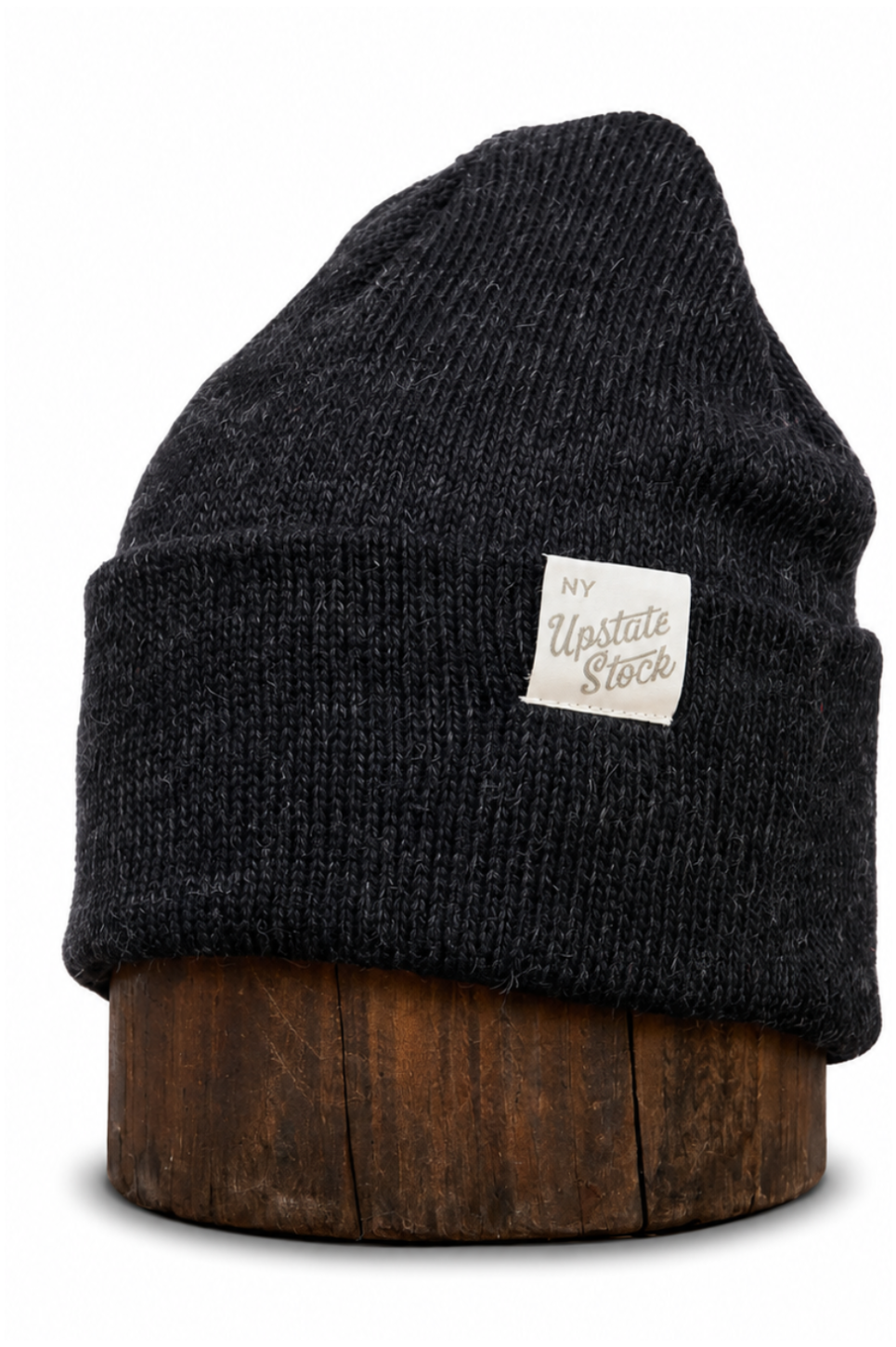 A dark charcoal gray ribbed knit beanie hat with a small white rectangular patch reading 'My Upstate Stock', displayed on a round wooden stand against a white background.