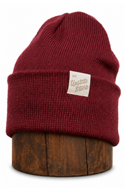 A dark red ribbed knit beanie hat displayed on a round wooden stand against a plain white background. The beanie features a small beige fabric label on the front cuff that reads 'NY Upstate Store'.