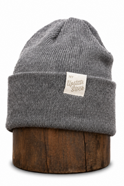 A gray ribbed knit beanie hat with a folded cuff and a small cream label reading 'My Upstate Stock,' displayed on a round wooden stand against a white background.