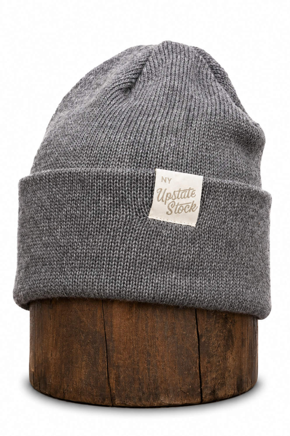A gray ribbed knit beanie hat with a folded cuff and a small cream label reading 'My Upstate Stock,' displayed on a round wooden stand against a white background.