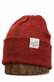 A rust red ribbed knit beanie hat with a small white rectangular 'My Yupoong Stock' label on the front, displayed on a round wooden riser against a white background.