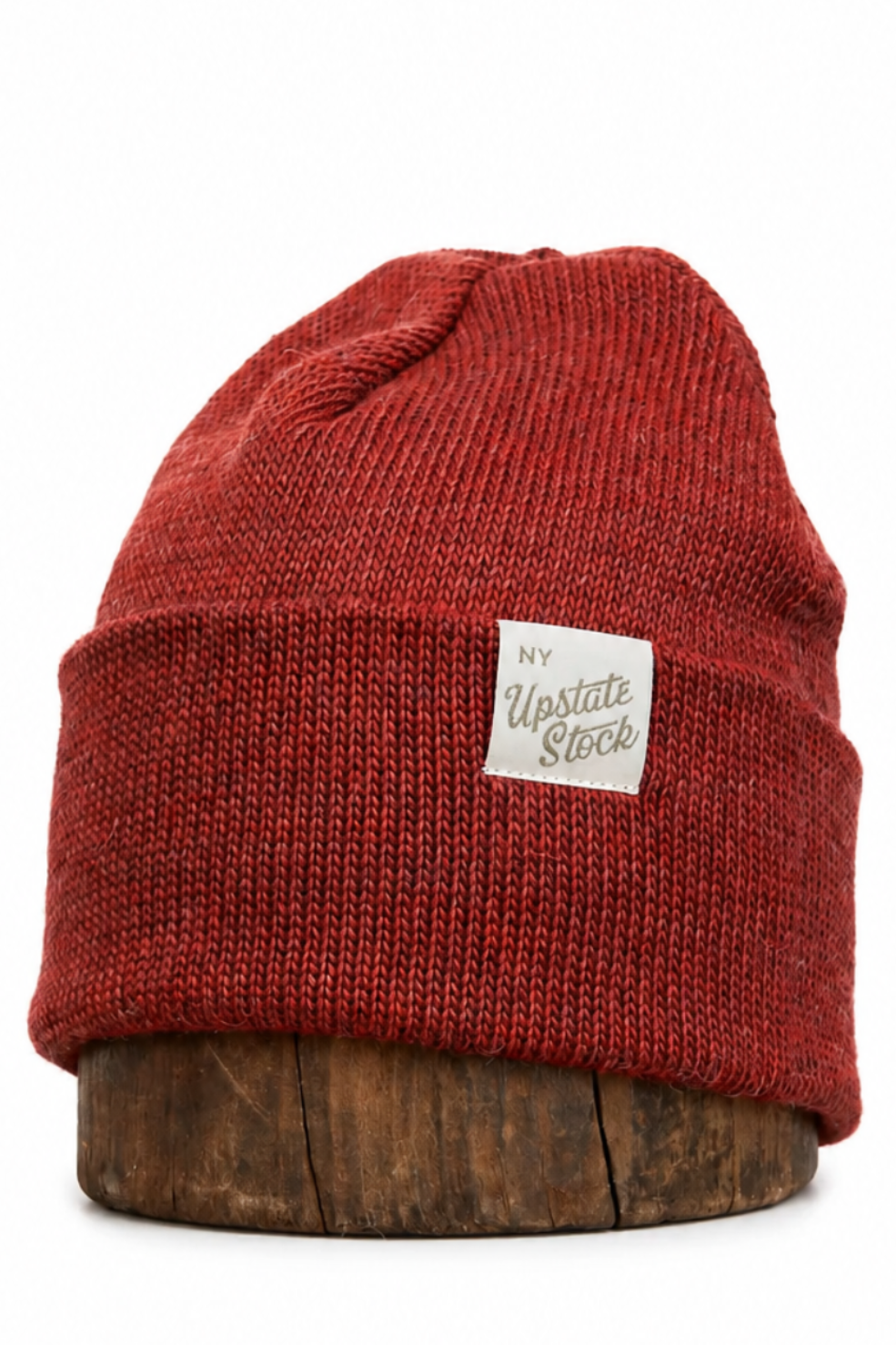 A rust red ribbed knit beanie hat with a small white rectangular 'My Yupoong Stock' label on the front, displayed on a round wooden riser against a white background.