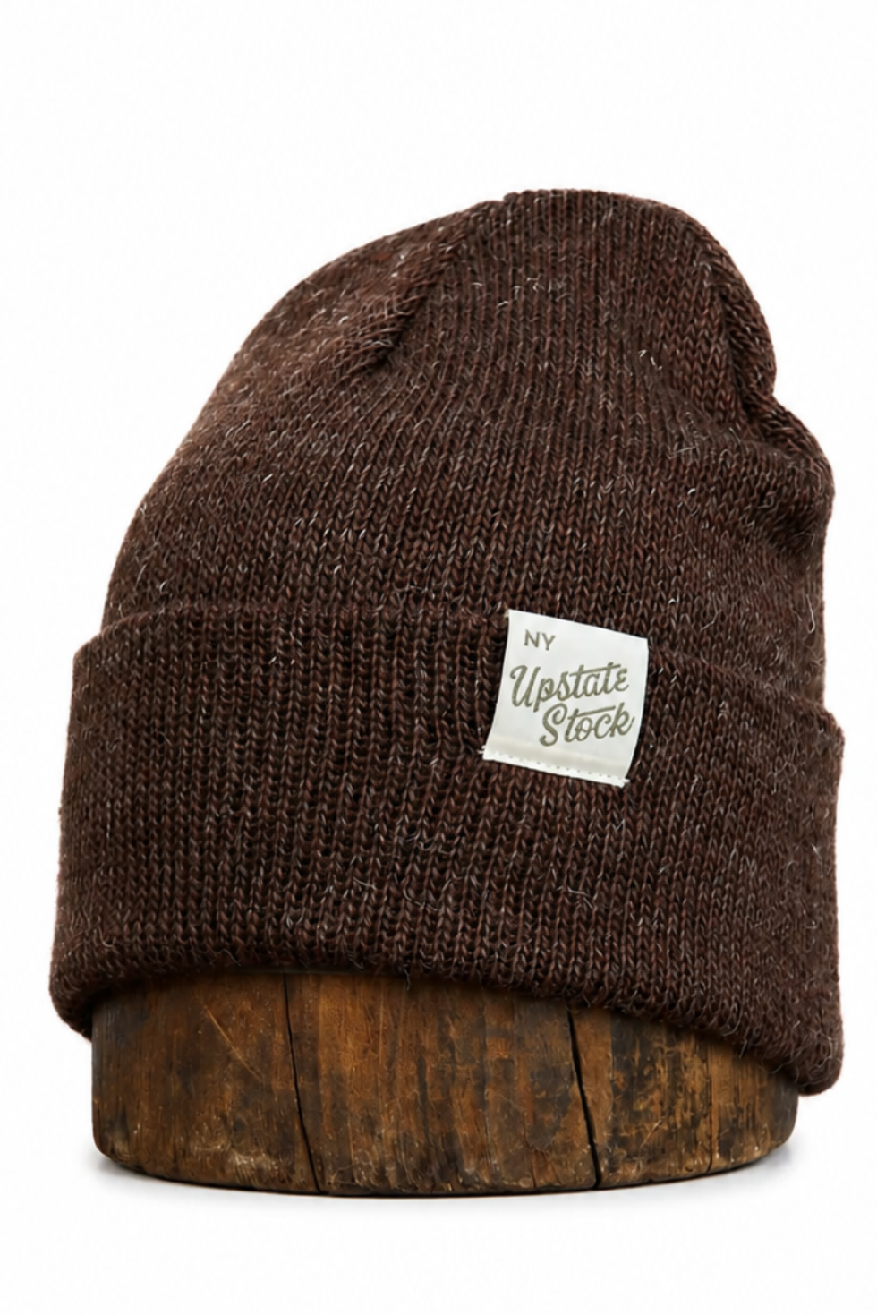 A dark brown ribbed knit beanie hat with a small white 'Upstate Stock' brand patch on the front cuff, displayed on a wooden stand against a plain white background.