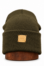 An olive green ribbed knit beanie hat with a tan leather patch on the front, displayed on a brown wooden stand against a white background.