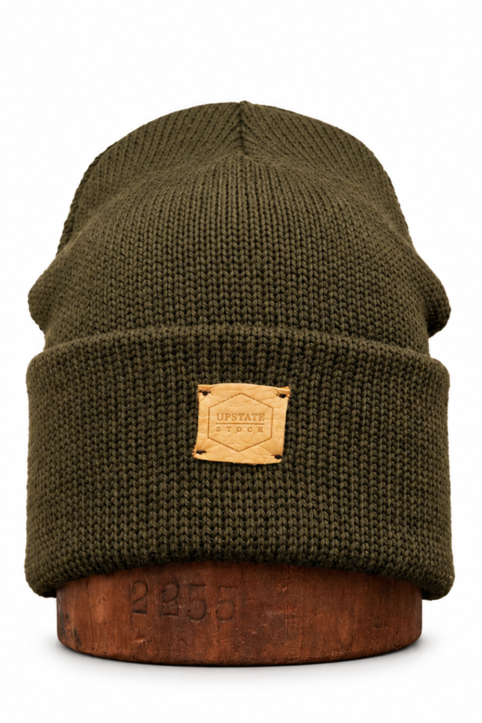 An olive green ribbed knit beanie hat with a tan leather patch on the front, displayed on a brown wooden stand against a white background.