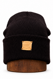 A black ribbed knit beanie hat with a tan leather patch on the front, displayed on a wooden stand against a white background.
