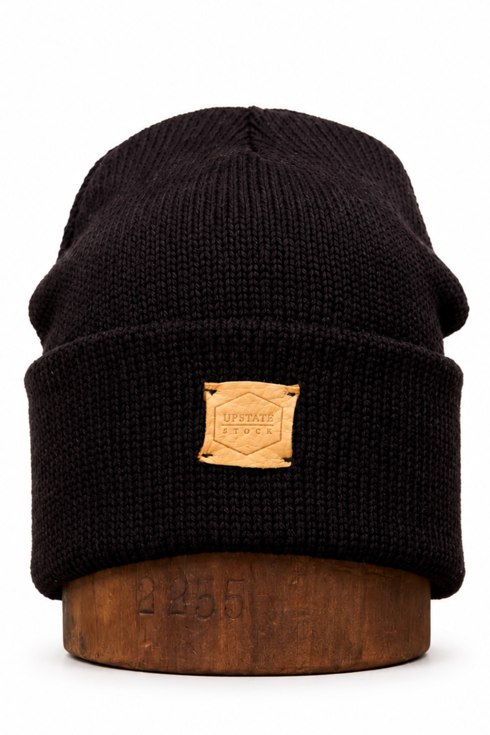 A black ribbed knit beanie hat with a tan leather patch on the front, displayed on a wooden stand against a white background.
